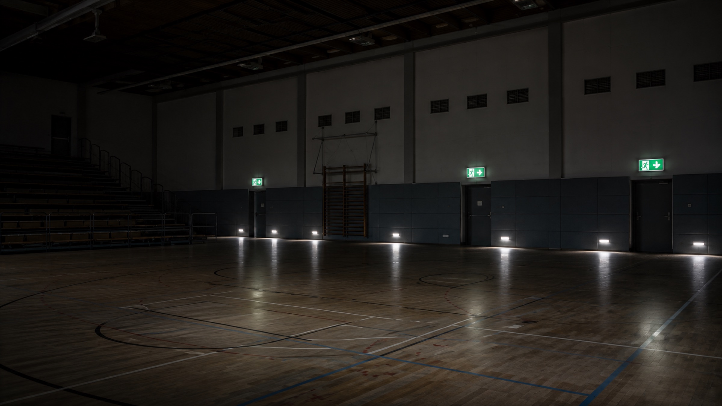 Unbeleuchtete Sporthalle, nur Rettungszeichen und bodennahe Sicherheitsleuchten an der Längswand